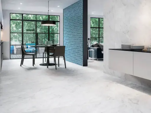 Modern open dining area with marble-look floor, round dining table with four chairs, blue brick accent wall and large windows with a view of greenery.