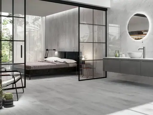 Stylish modern bedroom with adjoining bathroom, grey wood-look floor, glass sliding doors and a round illuminated mirror above the sink.